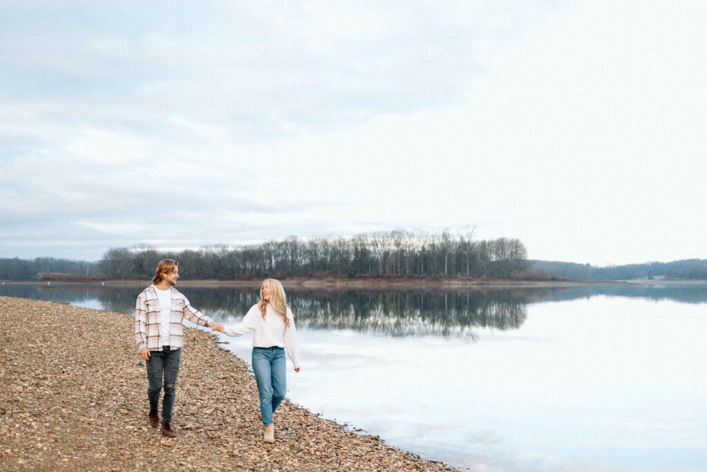 Engaged couple walking hand in hand along the shoreline of a large lake at Cordorus State Park. 