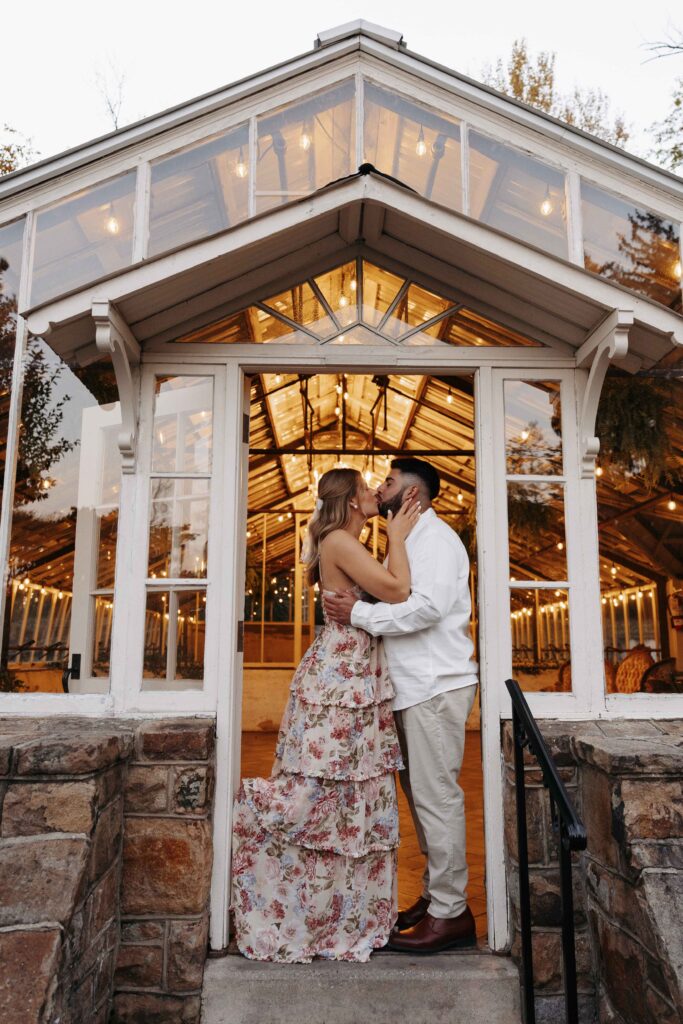 A couple kissing in the doorway of the greenhouse at Historic Shady Lane with twinkle lights captured by their engagement photographer. 