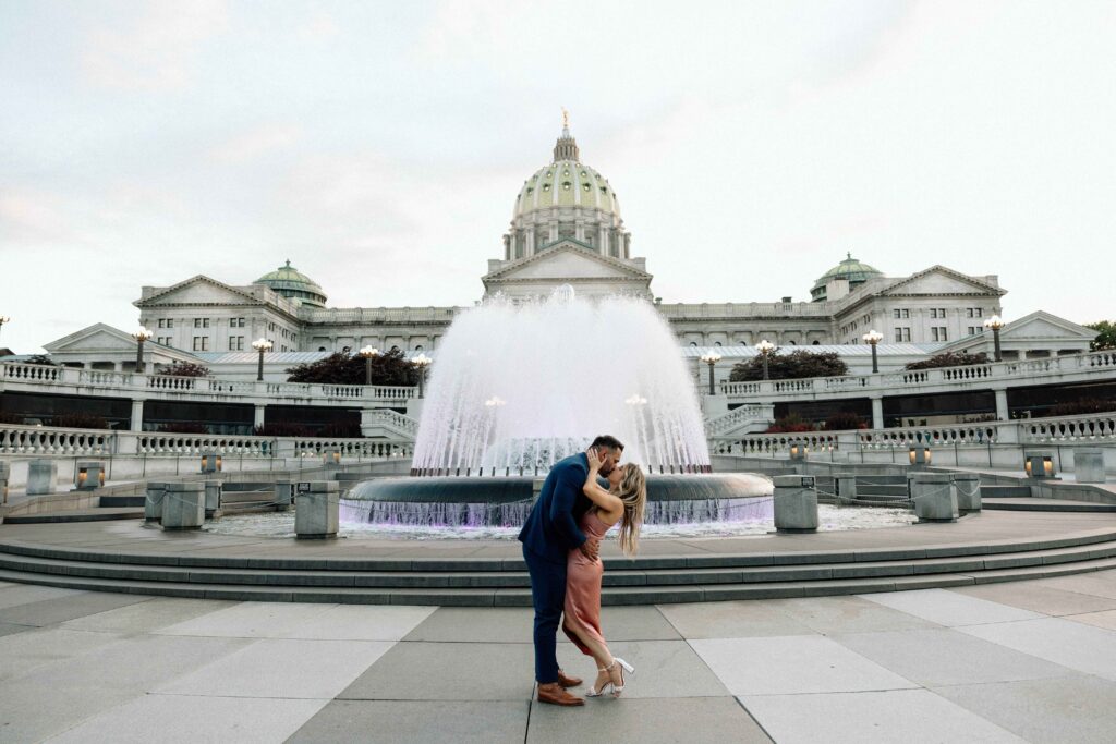 Couple kissing in front of the large fountain at the Pennsylvania State Capital captured by their engagement photographer. 