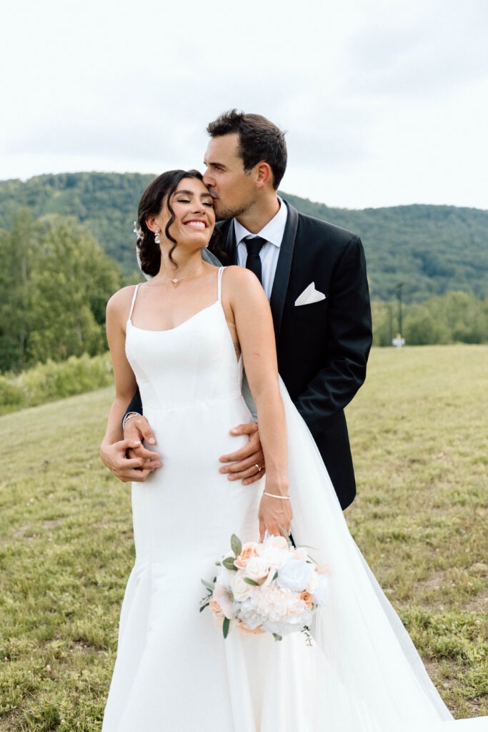 A bride smiling while the groom kisses her head. The bride is holding her bridal bouquet which is a wedding budget idea.