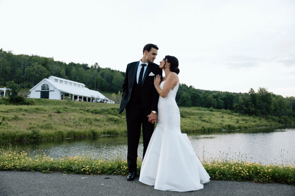 A newlywed couple posing in front of their wedding venue, a lake, and mountains.