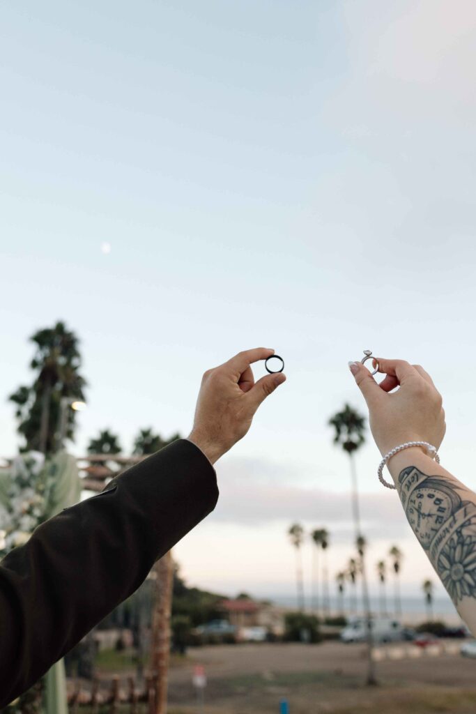 Bride and groom holding their wedding rings in the air with palm trees in the background.