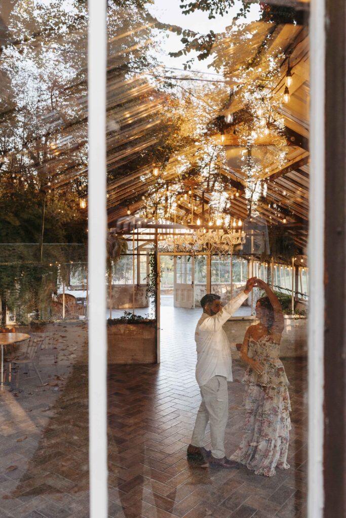 A couple dancing inside a greenhouse. The photo is taken from outside the window, looking in.