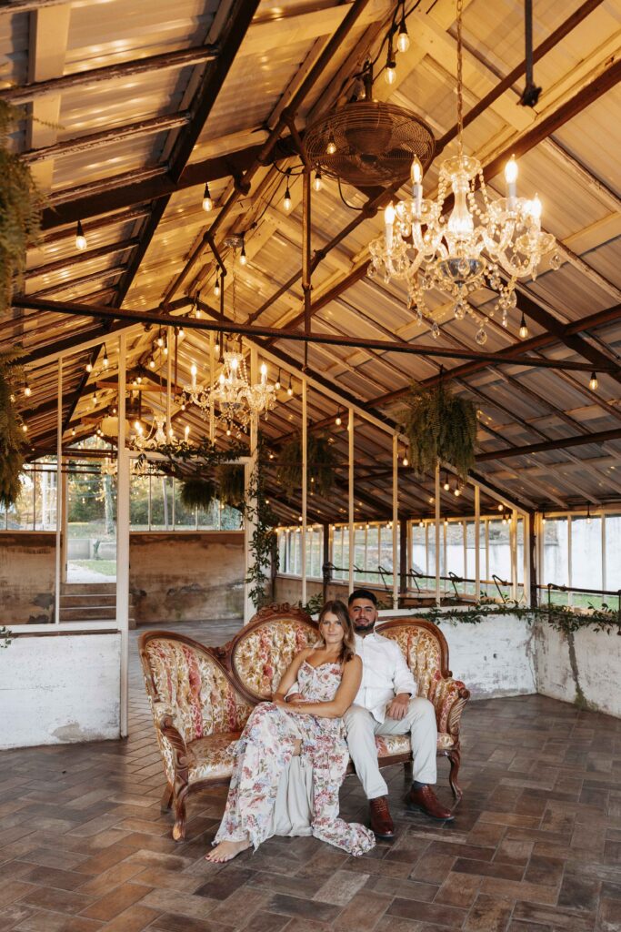 Couple sitting on a vintage couch inside a greenhouse full of warm light during their romantic engagement photoshoot.