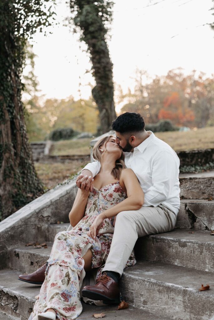 Couple sitting and kissing on the steps with tress in the background.