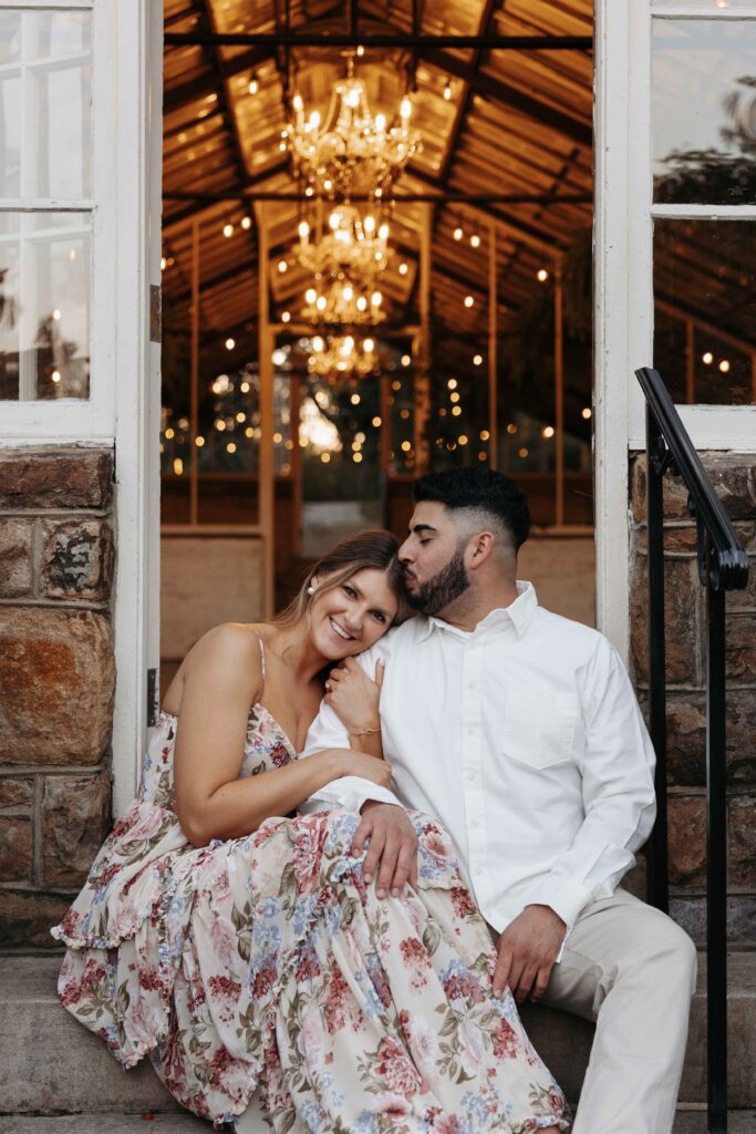 A couple sitting on the steps in front of a greenhouse with twinkle lights shining behind them.