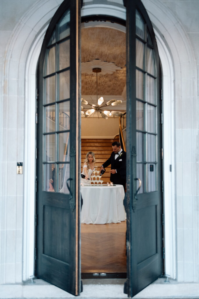 Elegant wedding photos of the bride and groom pouring champagne into their champagne tower, taken outside a doorway.