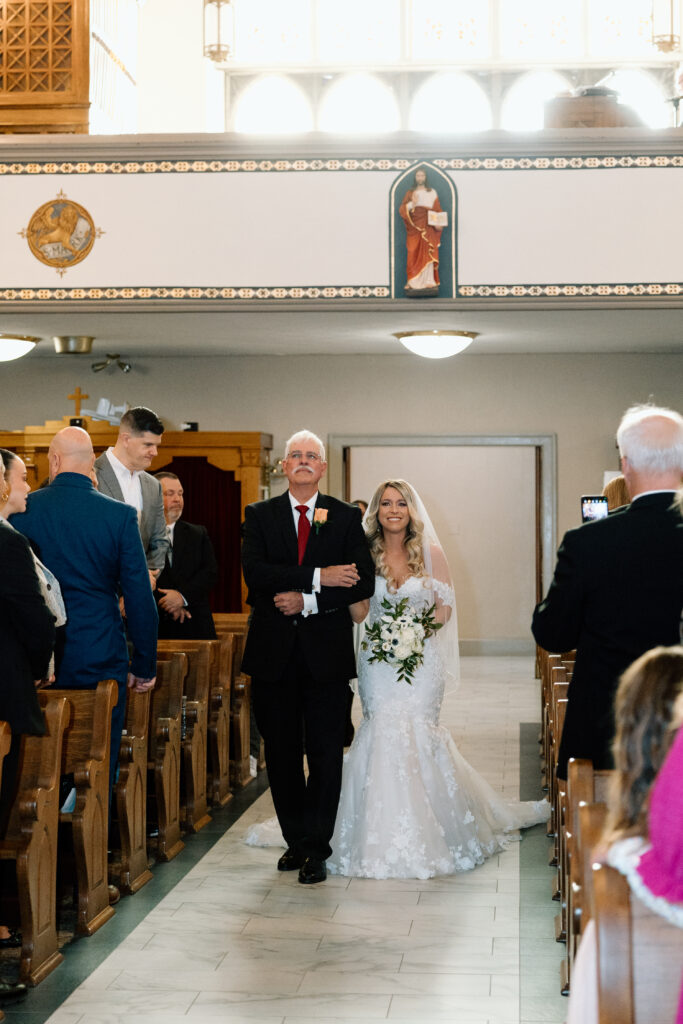 The bride walking down the aisle with her father and smiling at her groom. 