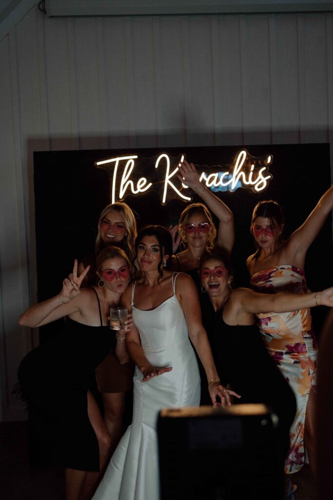 Bride and bridesmaids posing for a photo booth with a neon sign in the background. 