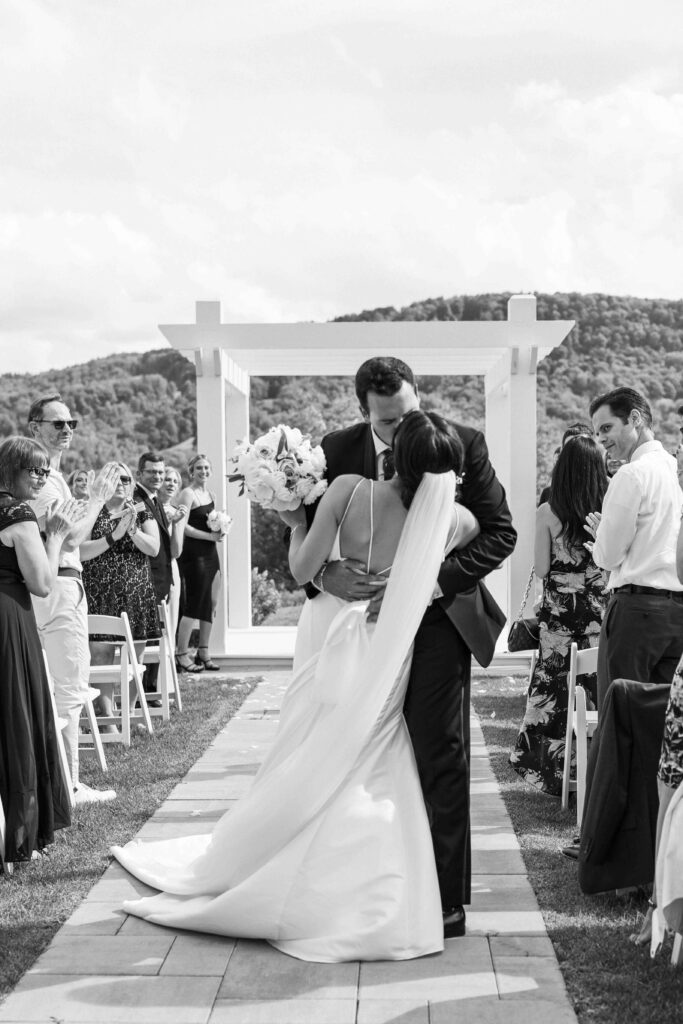 Bride and groom kissing at the end of the aisle while guests clap. 