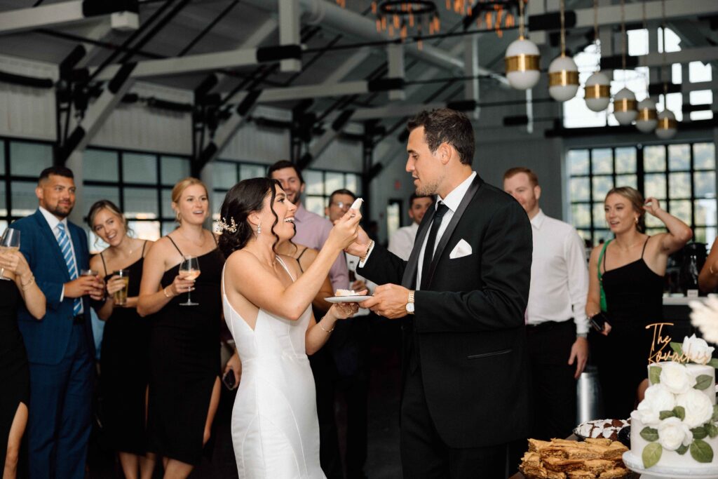 Bride and groom trying their wedding cake with guests smiling in the background at their black and white wedding.