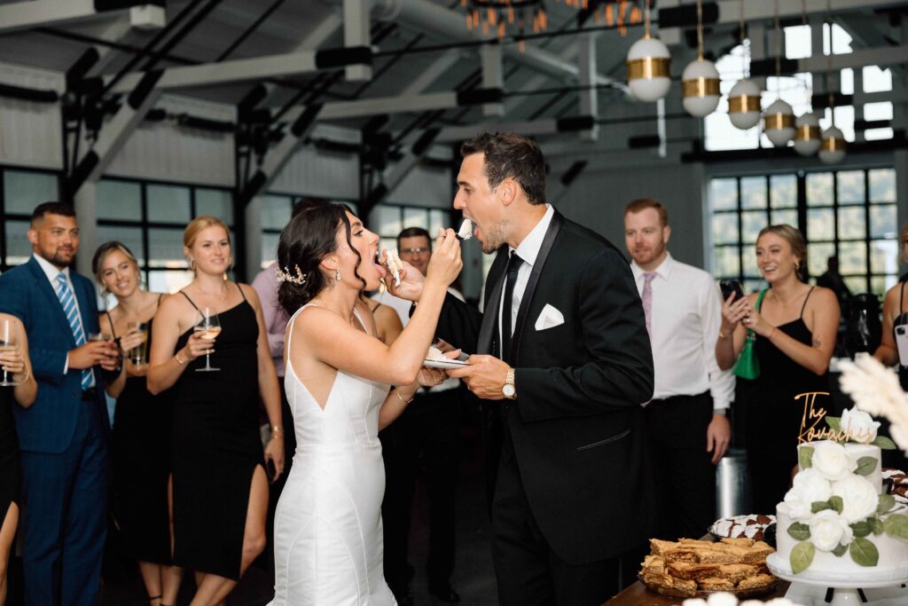 Bride and groom feeding each other cake as guests laugh. 