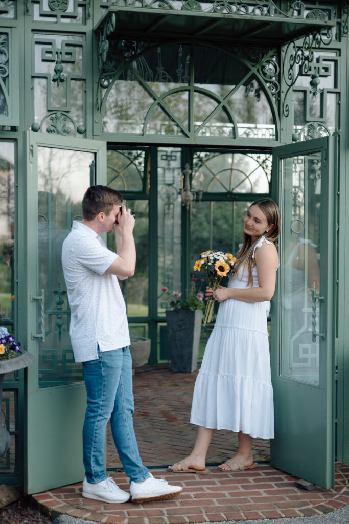 Engagement photo poses of a man taking a photo of his fiance holding flowers.