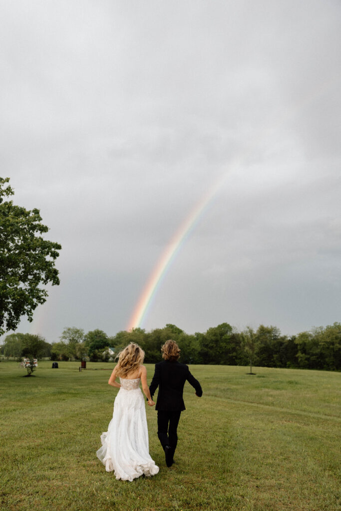 Editorial wedding photography of a bride and groom holding hands and running towards a rainbow. 