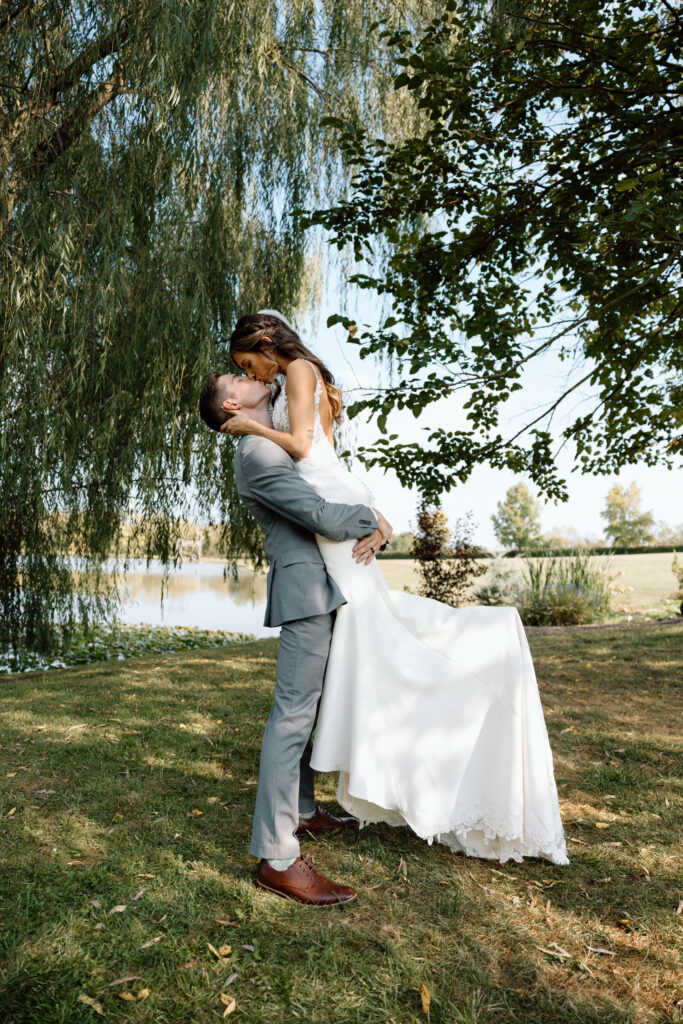 Groom picking up his wife and kissing romantically in front of a scenic lake and trees. 