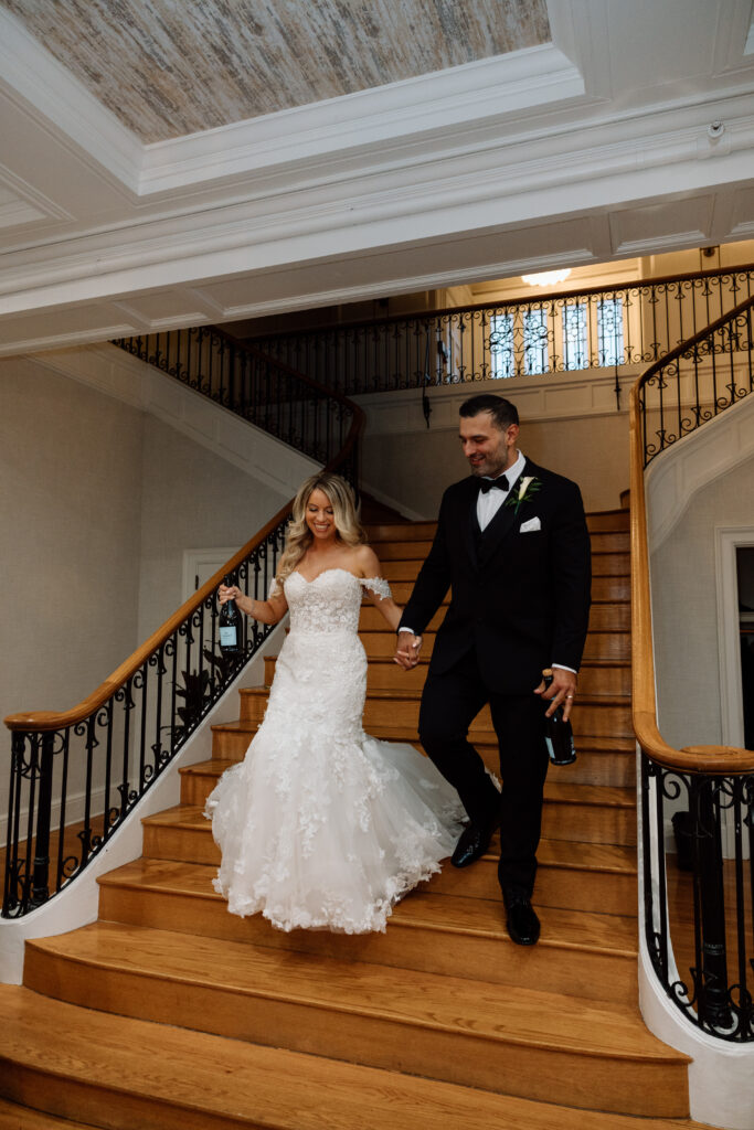 Bride and groom walking down the staircase at King Mansion holding champagne. 