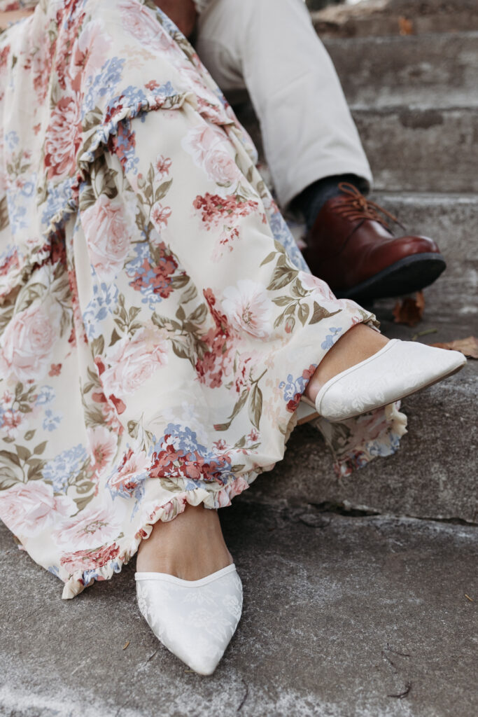 Close up of bride and grooms engagement session outfits, floral dress, white heels, dress pants, and dress shoes. 